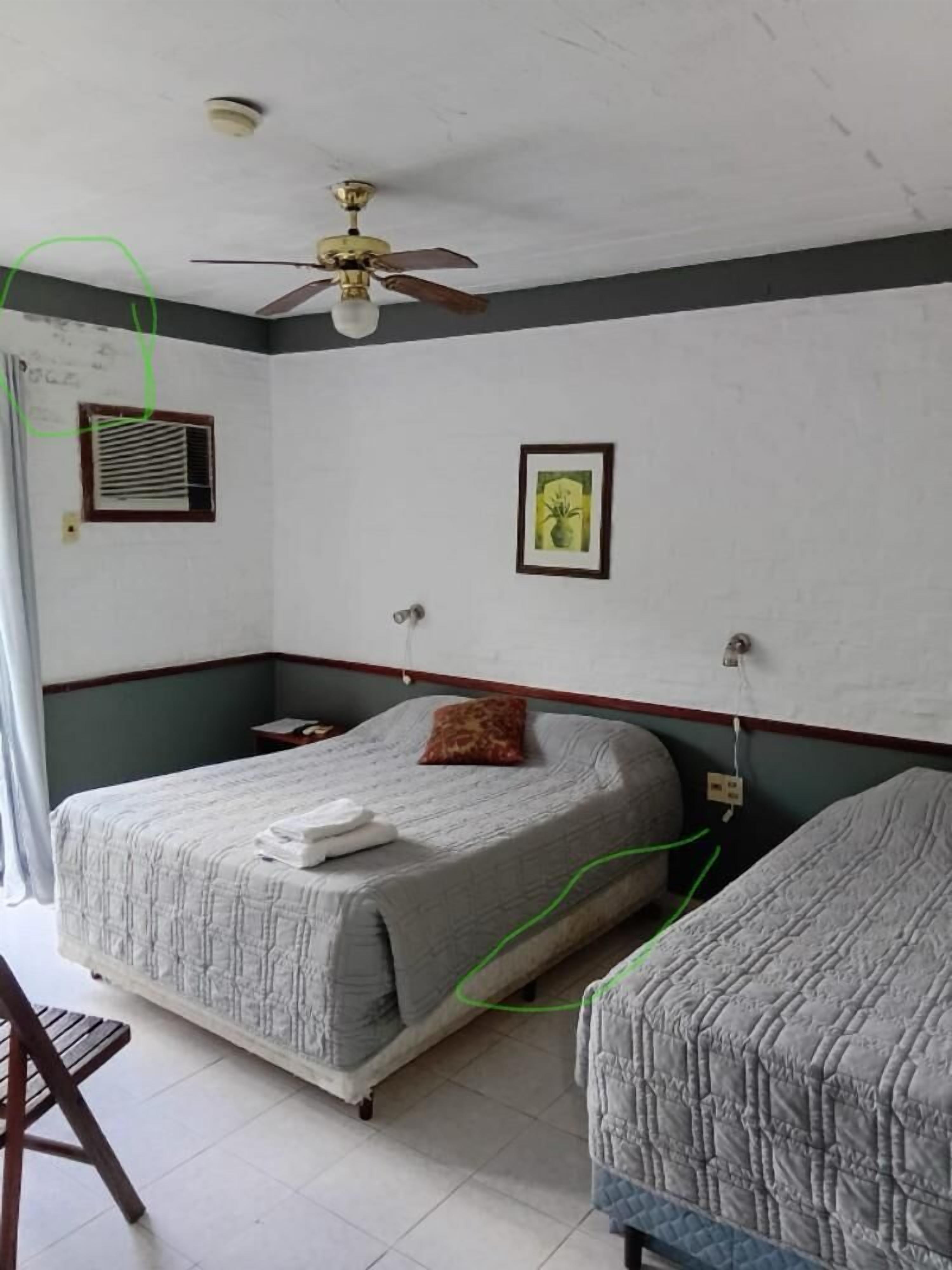 A hotel room with a bed, a bedside table, a ceiling fan, a window with curtains, and a wall-mounted air conditioning unit.