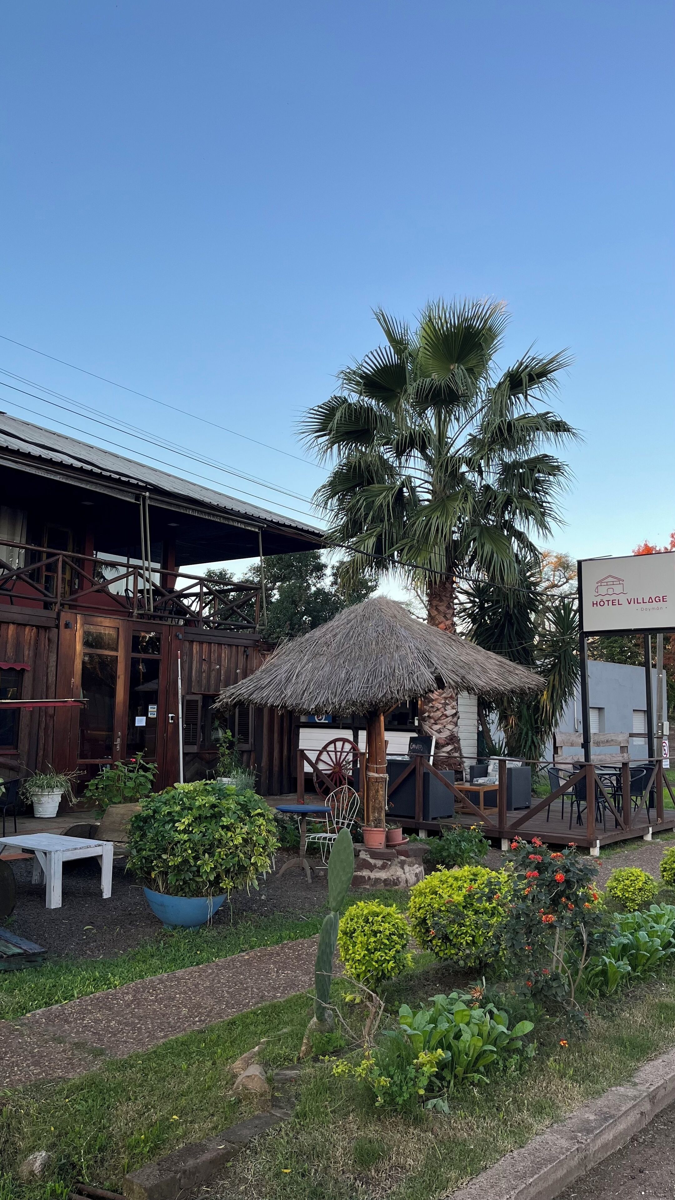 A rustic building with a thatched-roof outdoor seating area, surrounded by lush greenery and a palm tree.