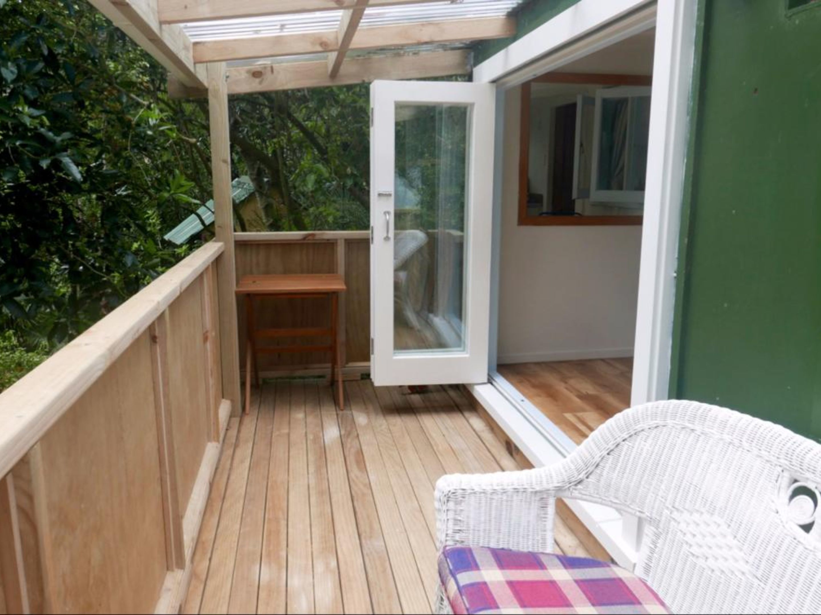 A wooden deck with a white wicker chair and a small table, a glass door leading to a room, and a green wall.
