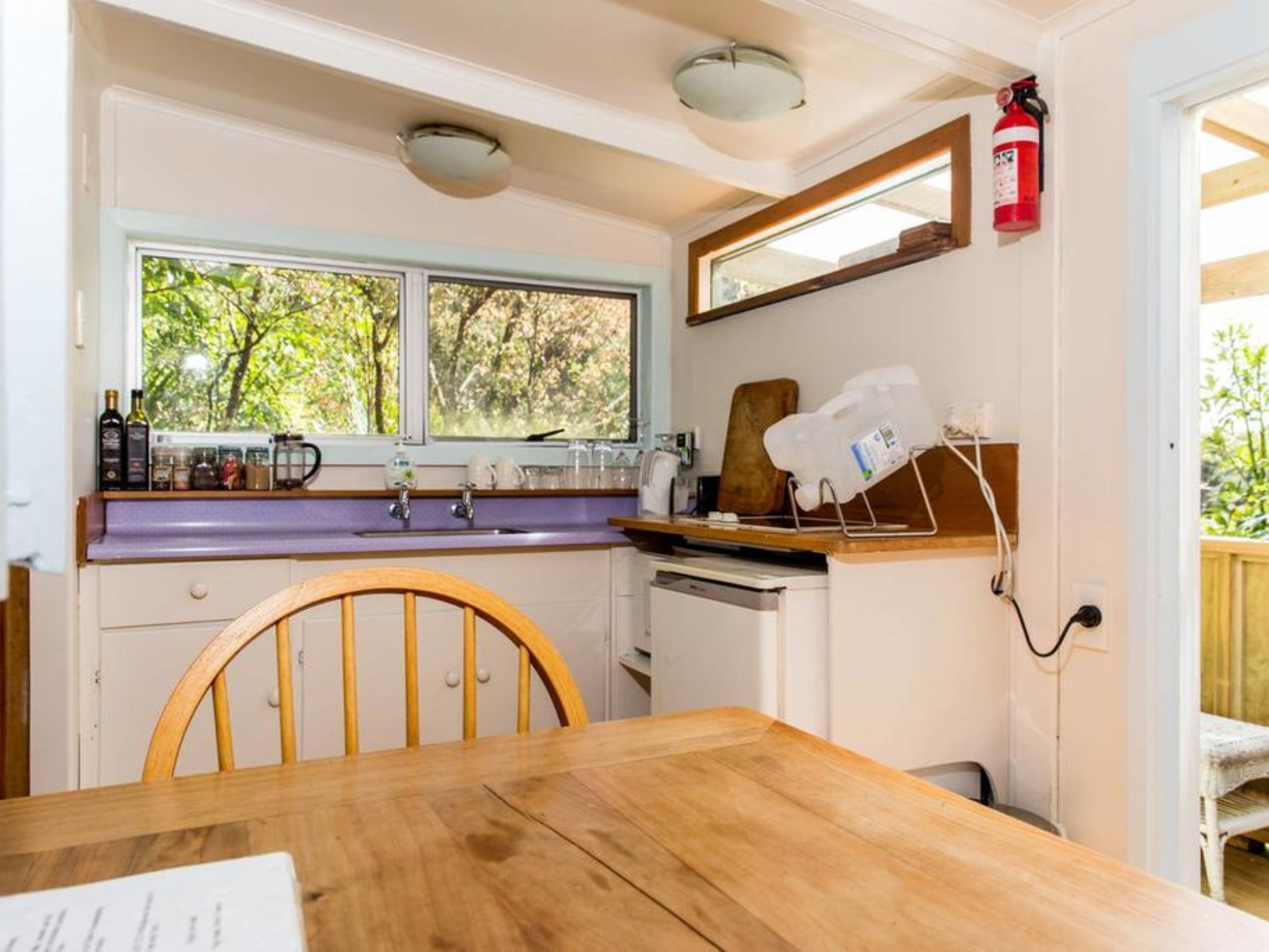A compact kitchen with a wooden table, a window with a view of greenery, a fire extinguisher, and a countertop with various items.