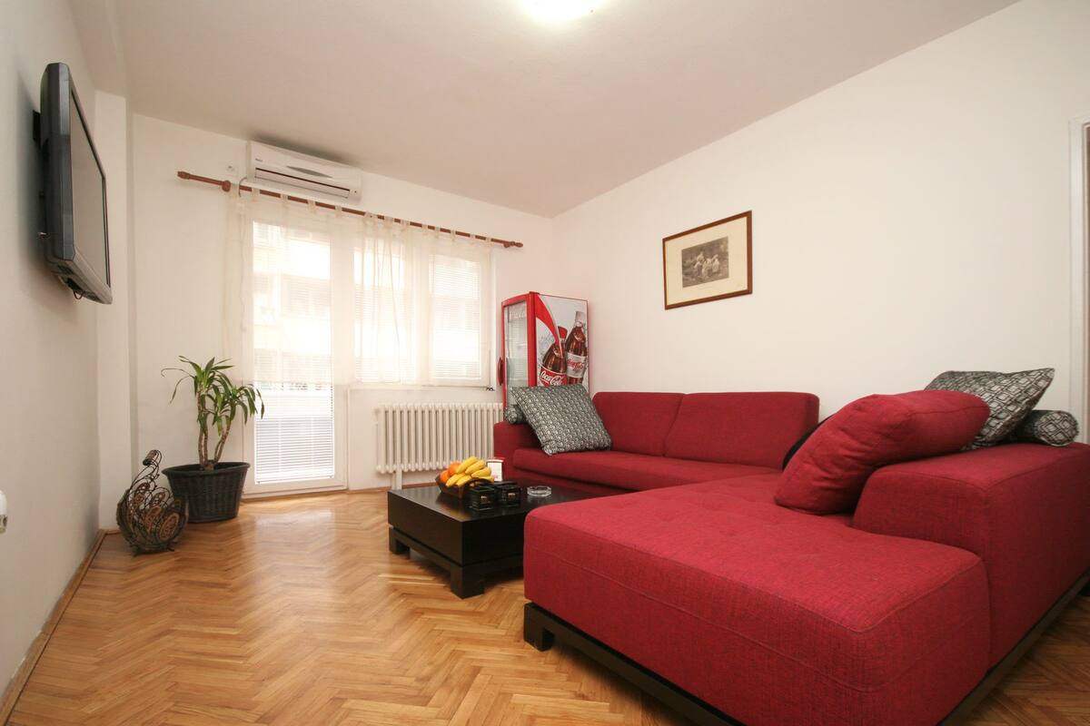 Una sala de estar con un sofá rojo, una mesa de centro negra con frutas, un radiador y una ventana con cortinas.