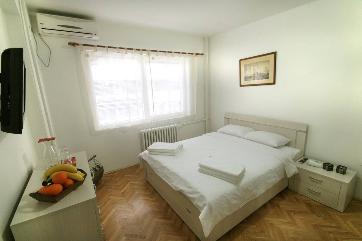 Un dormitorio con cama, mesita de noche, radiador, ventana con cortinas, aire acondicionado de pared y un cuadro enmarcado.
