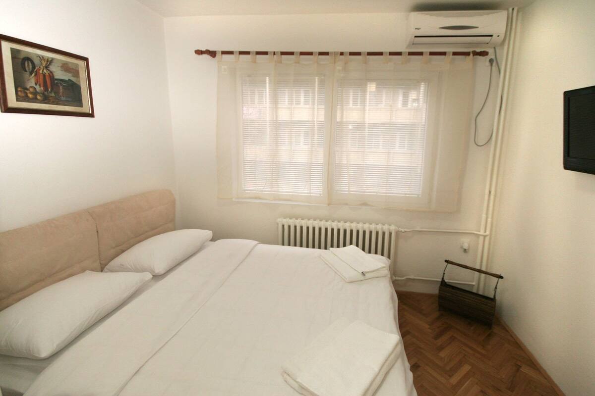 Un dormitorio con cama, radiador, ventana con persianas, equipo de aire acondicionado y un cuadro en la pared.