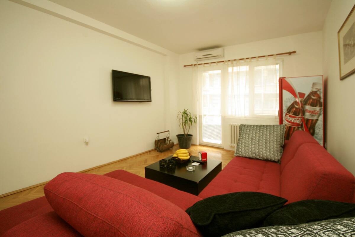 Una sala de estar con un sofá rojo, una mesa de centro negra, una planta en maceta y un televisor en la pared.