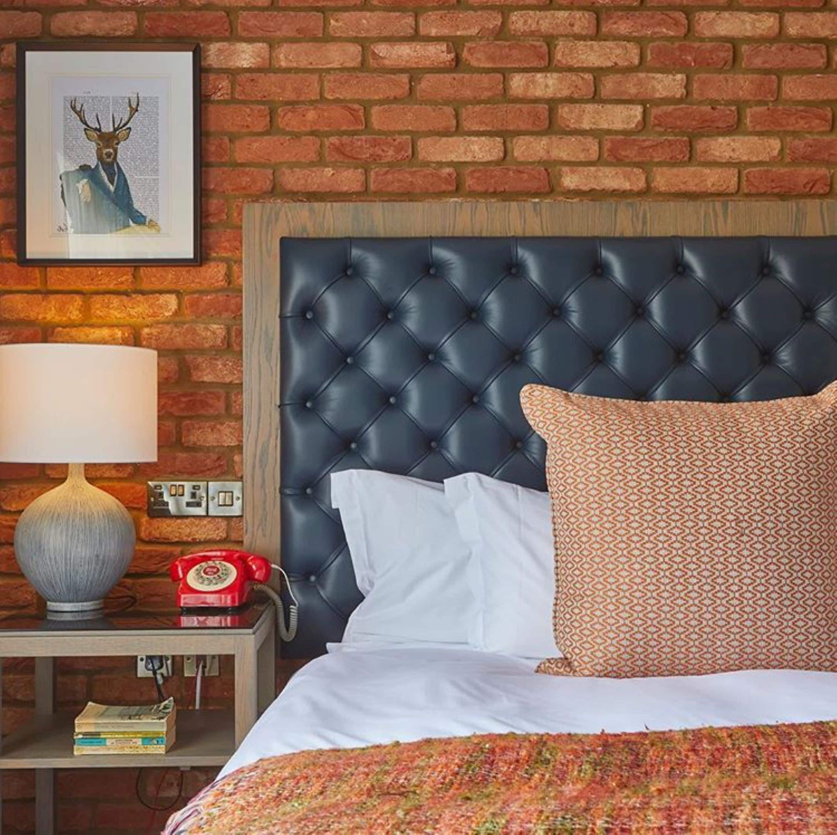 A hotel room with a brick wall, a tufted headboard, a patterned pillow, a white bedspread, a red rotary phone, a lamp, and a framed picture on the wall.