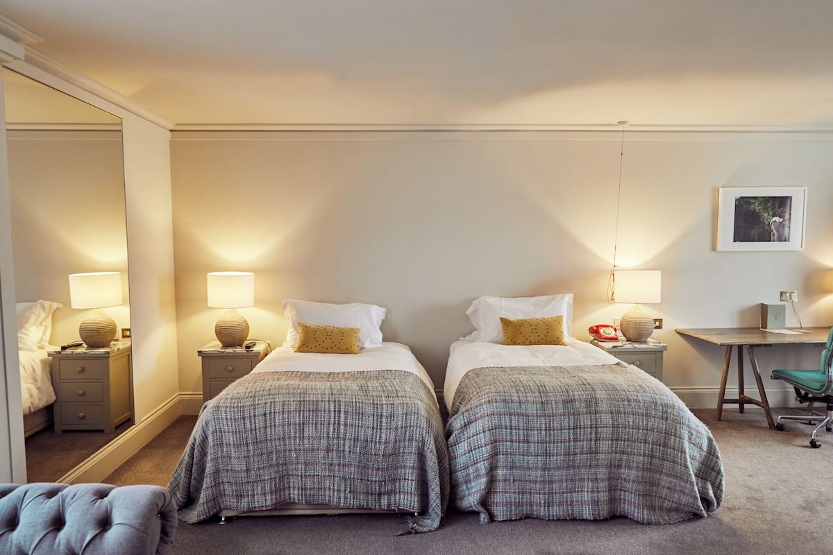 Habitación de hotel con dos camas, cada una con una colcha a cuadros grises y un almohadón amarillo. Hay lámparas de noche, un escritorio con una silla y un cuadro en la pared.