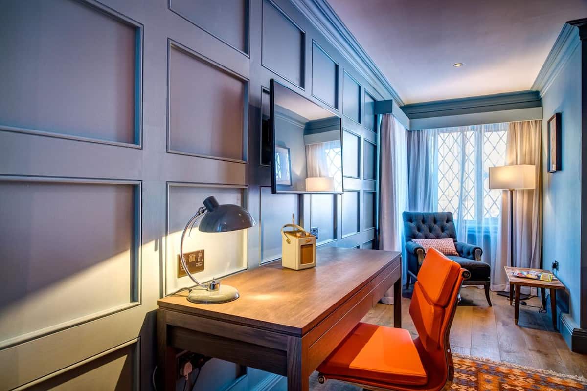 Habitación de hotel con escritorio de madera, una silla, una lámpara y un ventanal con cortinas.