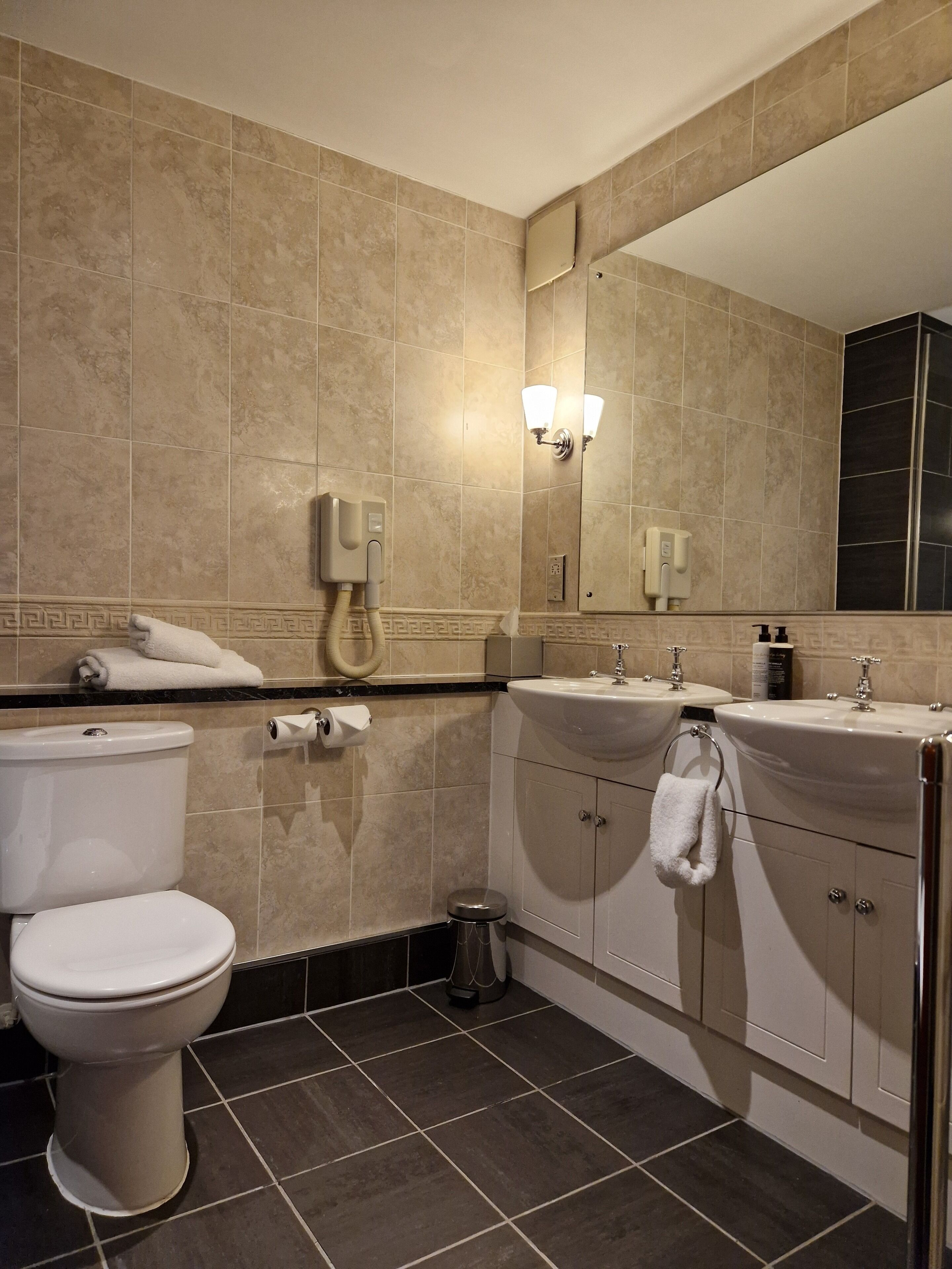 Baño con inodoro, lavamanos y espejo. Hay toallas en la encimera y un cubo de basura.