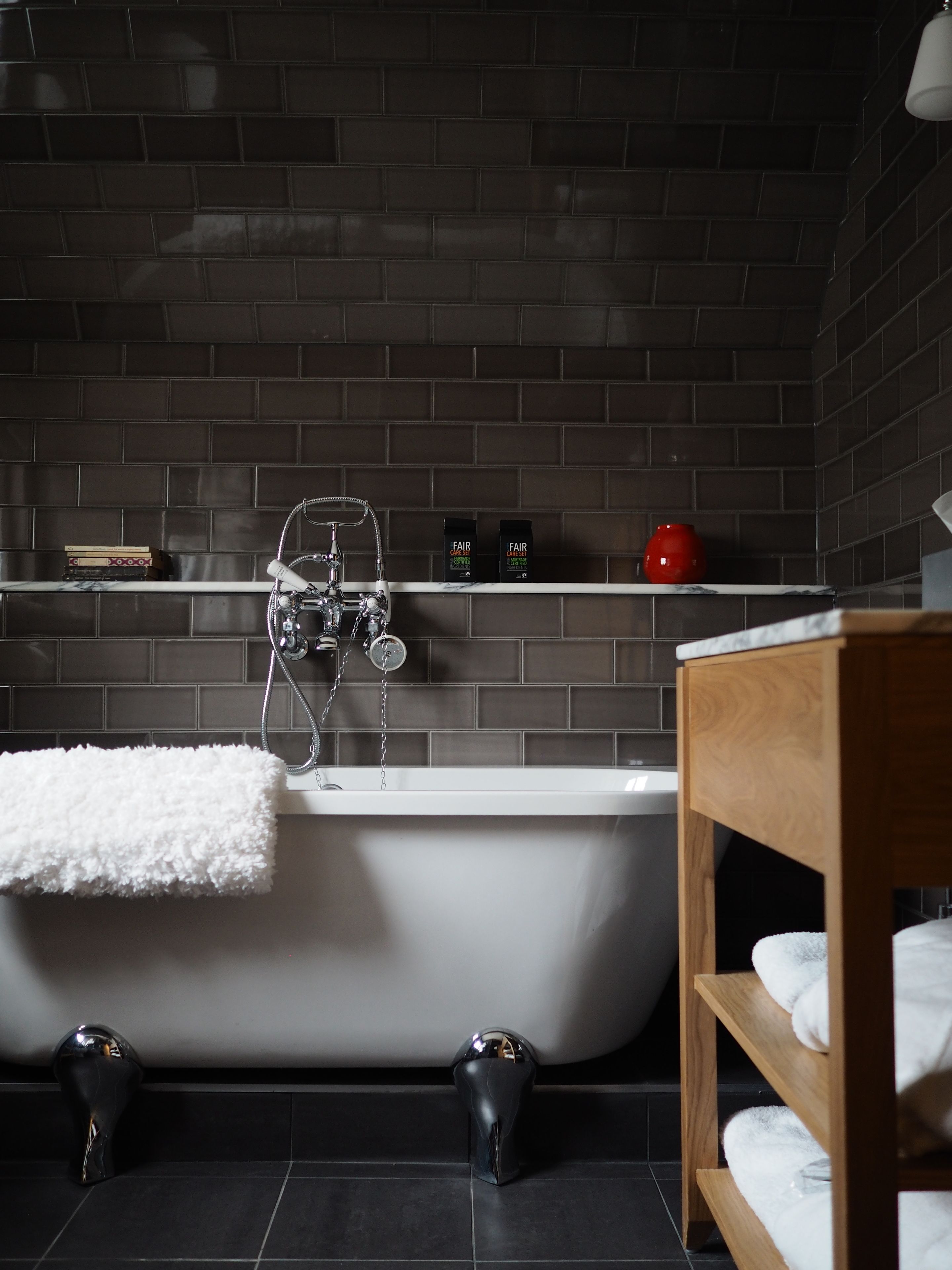 A modern bathroom with a freestanding bathtub, dark tiled walls, and a wooden shelf with toiletries.