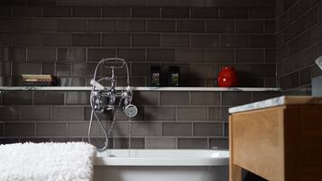 A modern bathroom with a freestanding bathtub, dark tiled walls, and a wooden shelf with toiletries.