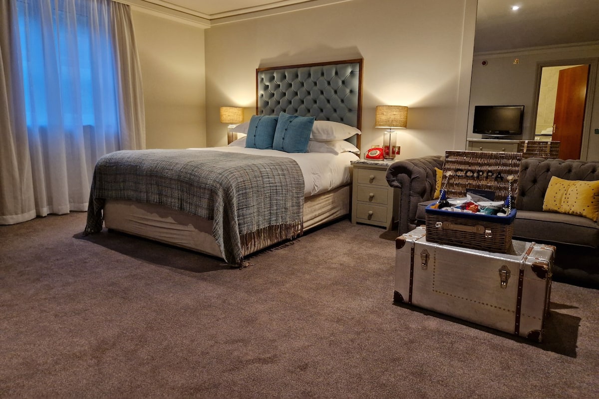 Habitación de hotel con una cama grande, una mesita de noche con lámpara, televisión, sofá y una maleta.
