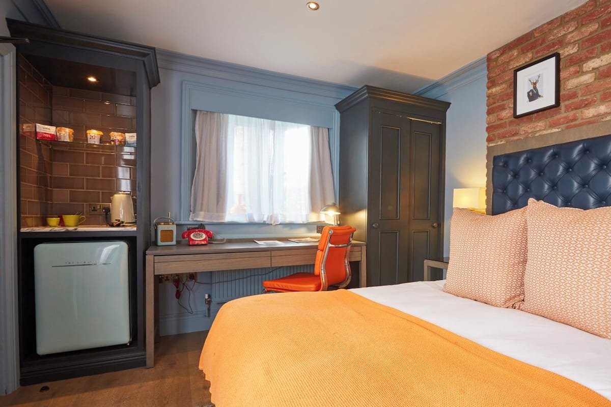 Habitación de hotel con cama, escritorio, silla, pequeña cocina y pared de ladrillo con cuadros.