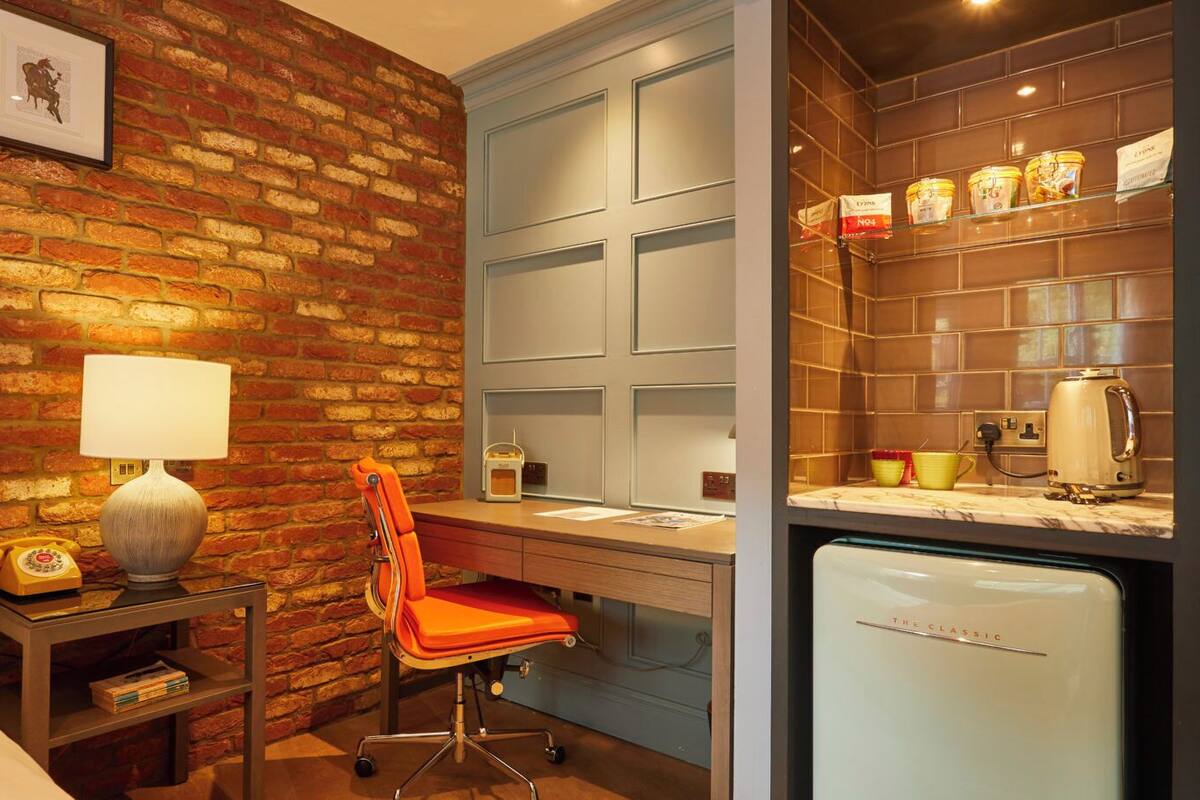 Habitación de hotel con pared de ladrillo, un escritorio con silla, una cama, una lámpara, un teléfono, un refrigerador y una estantería con frascos.