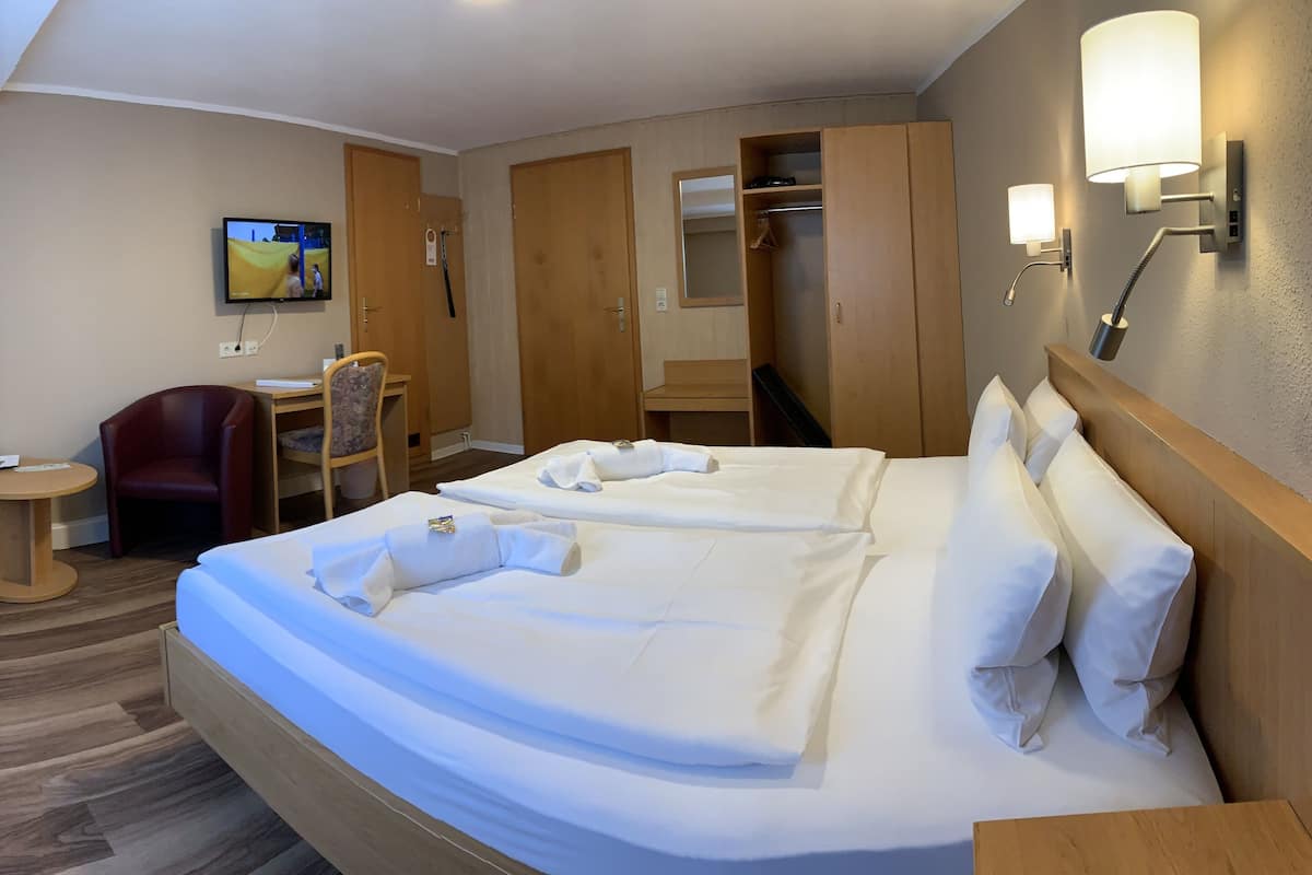 A hotel room with a large bed, a desk, a chair, a TV, and a wardrobe.