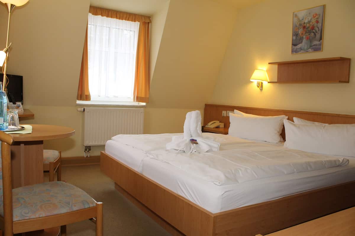 A hotel room with a bed, a desk, a chair, a lamp, a painting, and a window with curtains.