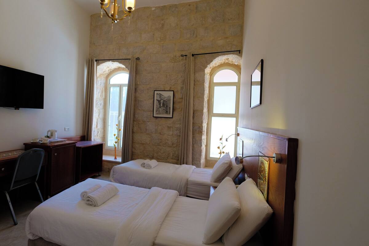 Quarto de hotel com duas camas, uma escrivaninha, uma cadeira, televisão e parede de pedra.