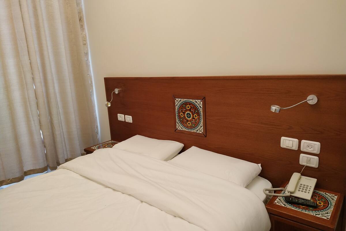 Cama bem arrumada com lençóis brancos, cabeceira de madeira, um pendurar decorativo na parede, um telefone ao lado da cama e uma mesinha de cabeceira com um jogo americano estampado.