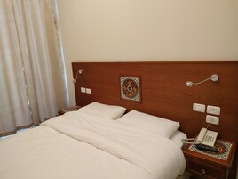 Una cama bien tendida con sábanas blancas, un cabecero de madera, un adorno en la pared, un teléfono en la mesita de noche y una mesita con un mantel individual estampado.