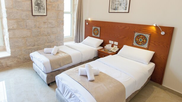 A hotel room with two beds, each with white linens and a wooden headboard. There are two bedside tables with lamps and a telephone. The room has stone walls and a window with curtains. - Azzahra Boutique Hotel & Restaurant - Jerusalem (Jerusalem)