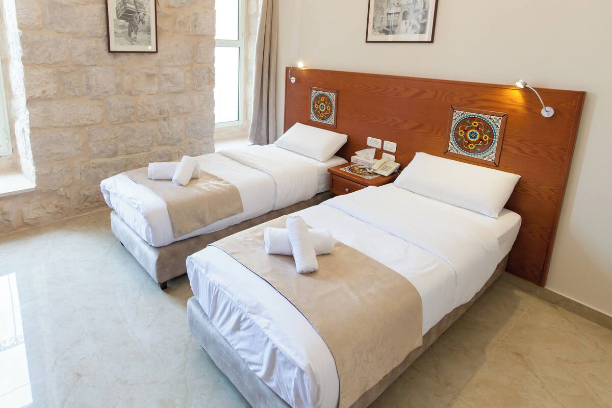 Quarto de hotel com duas camas, cada uma com roupa de cama branca e cabeceira de madeira. Há duas mesas de cabeceira com luminárias e telefone. O quarto possui paredes de pedra e uma janela com cortinas.