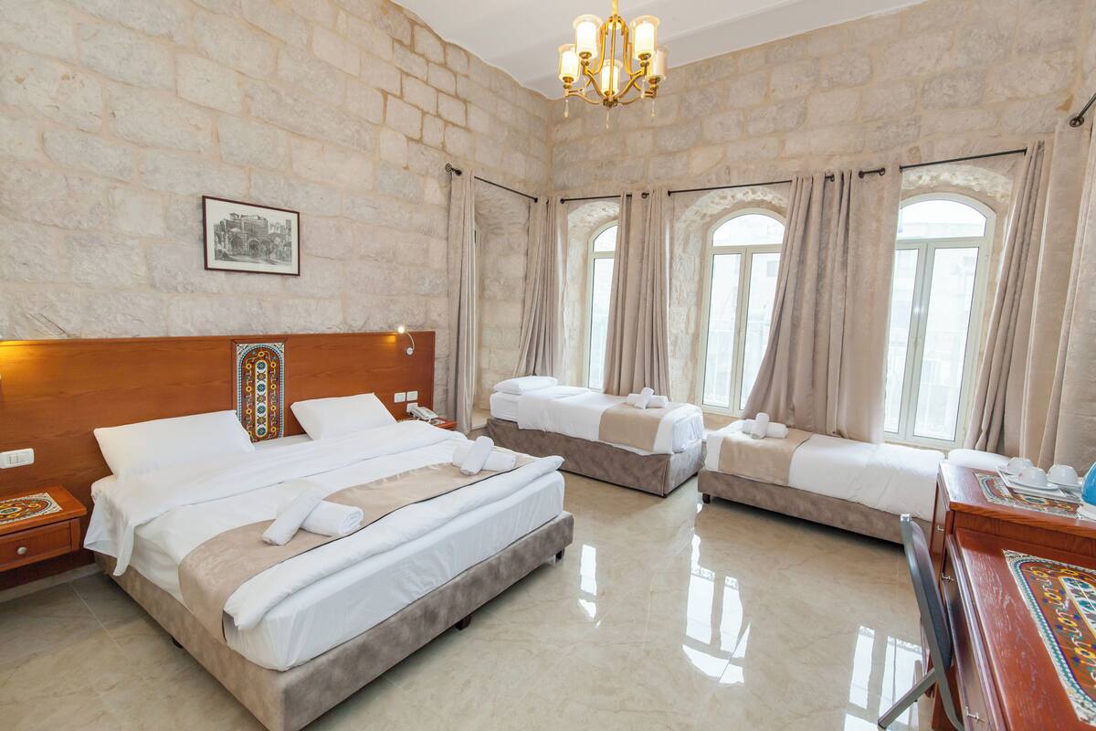 Quarto de hotel com três camas, cabeceira de madeira, um candelabro e janelas amplas com cortinas.