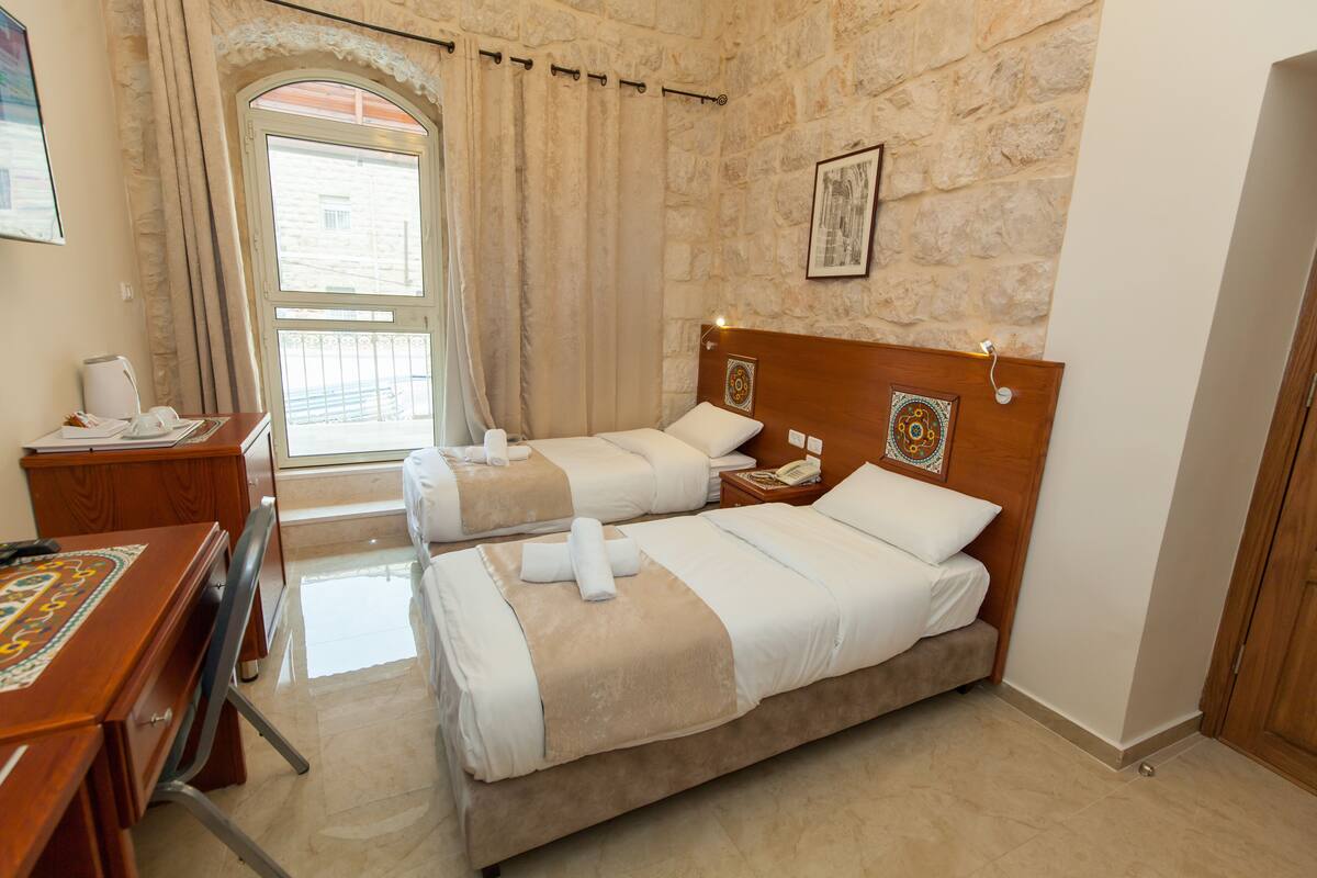 Quarto de hotel com duas camas, uma escrivaninha, uma cadeira, telefone e janela com cortinas.