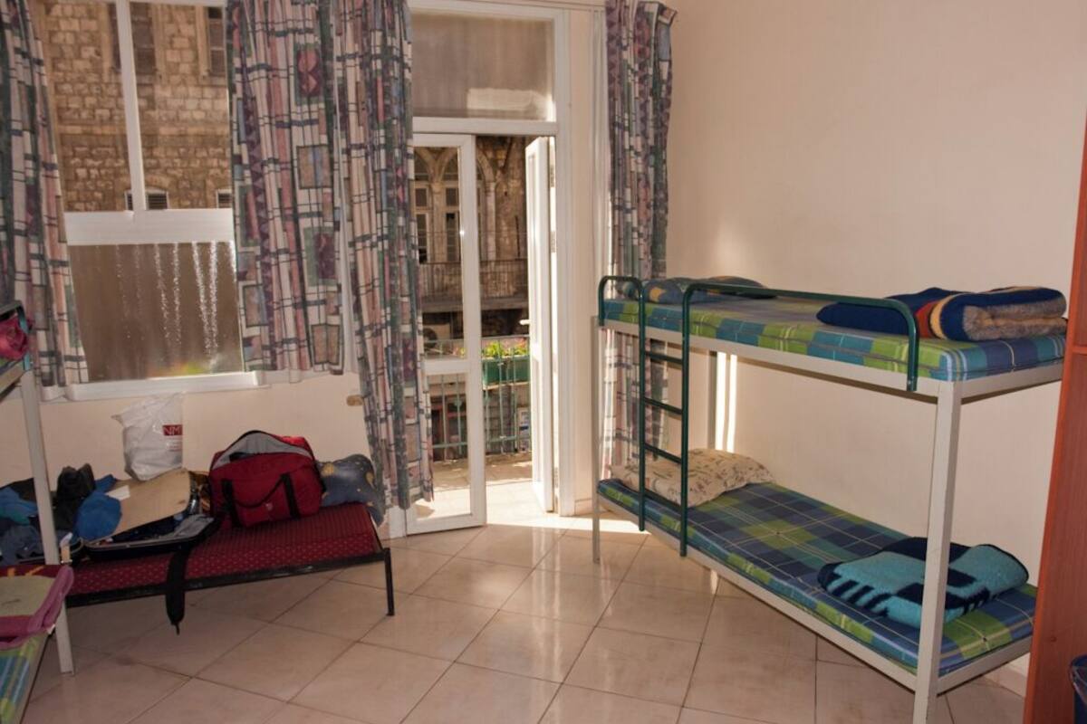 Um quarto de dormitório com beliches, uma janela com cortinas estampadas, uma mala e uma bolsa no chão.
