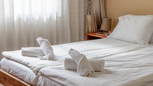 A neatly made bed with white linens and towels, a wooden nightstand with a lamp, and sheer curtains allowing natural light.