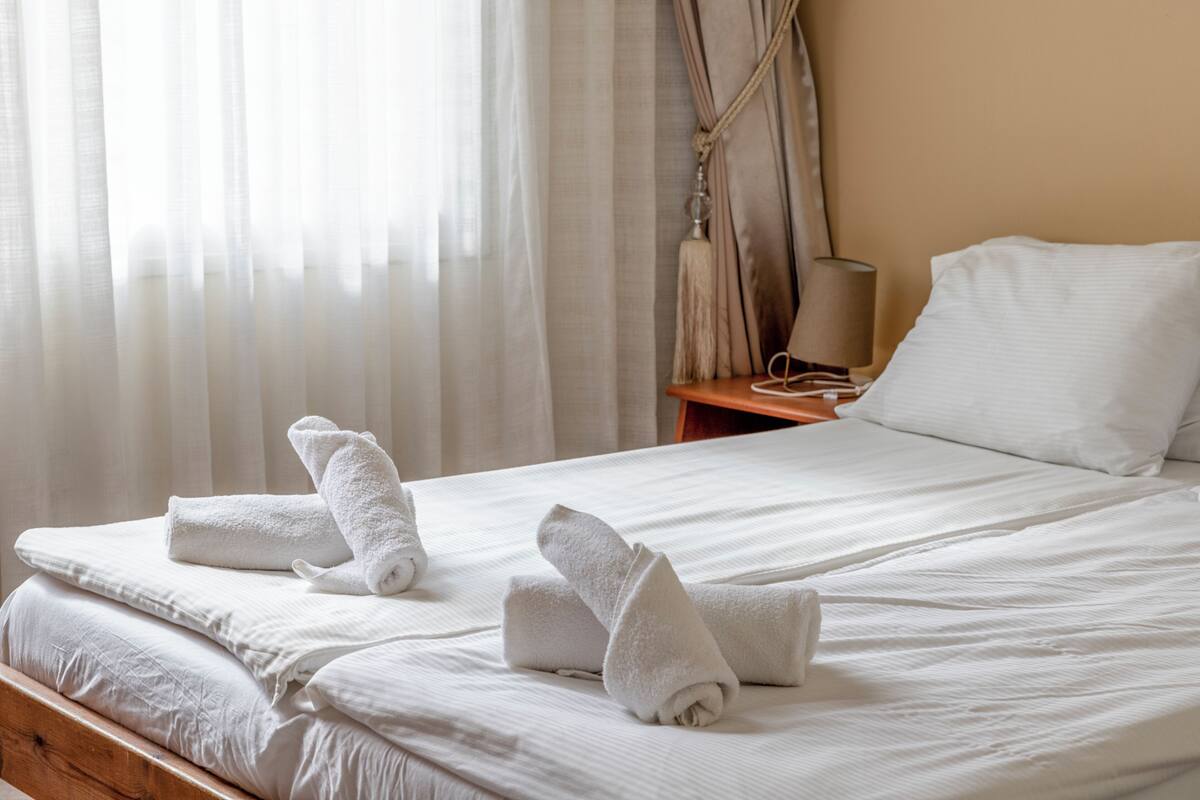 Cama bem arrumada com lençóis e toalhas brancos, mesa de cabeceira de madeira com abajur e cortinas leves que permitem a entrada de luz natural.