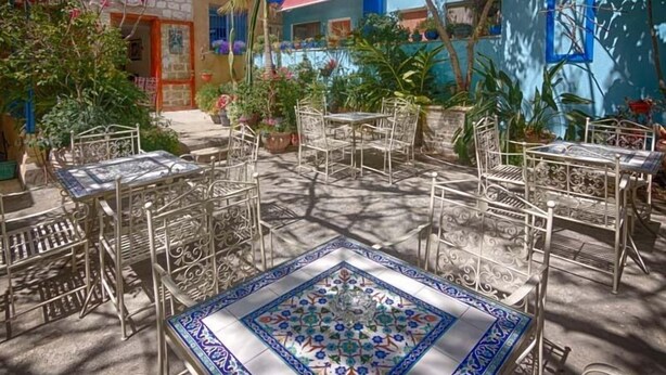 An outdoor patio with metal tables and chairs, surrounded by potted plants and a colorful building in the background.