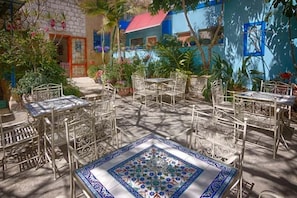 An outdoor patio with metal tables and chairs, surrounded by potted plants and a colorful building in the background.