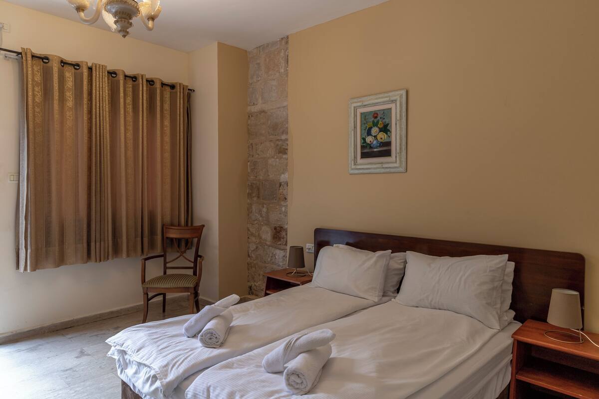 Quarto de hotel com cama de casal, uma cadeira, mesa de cabeceira, luminária, um quadro na parede e cortina.