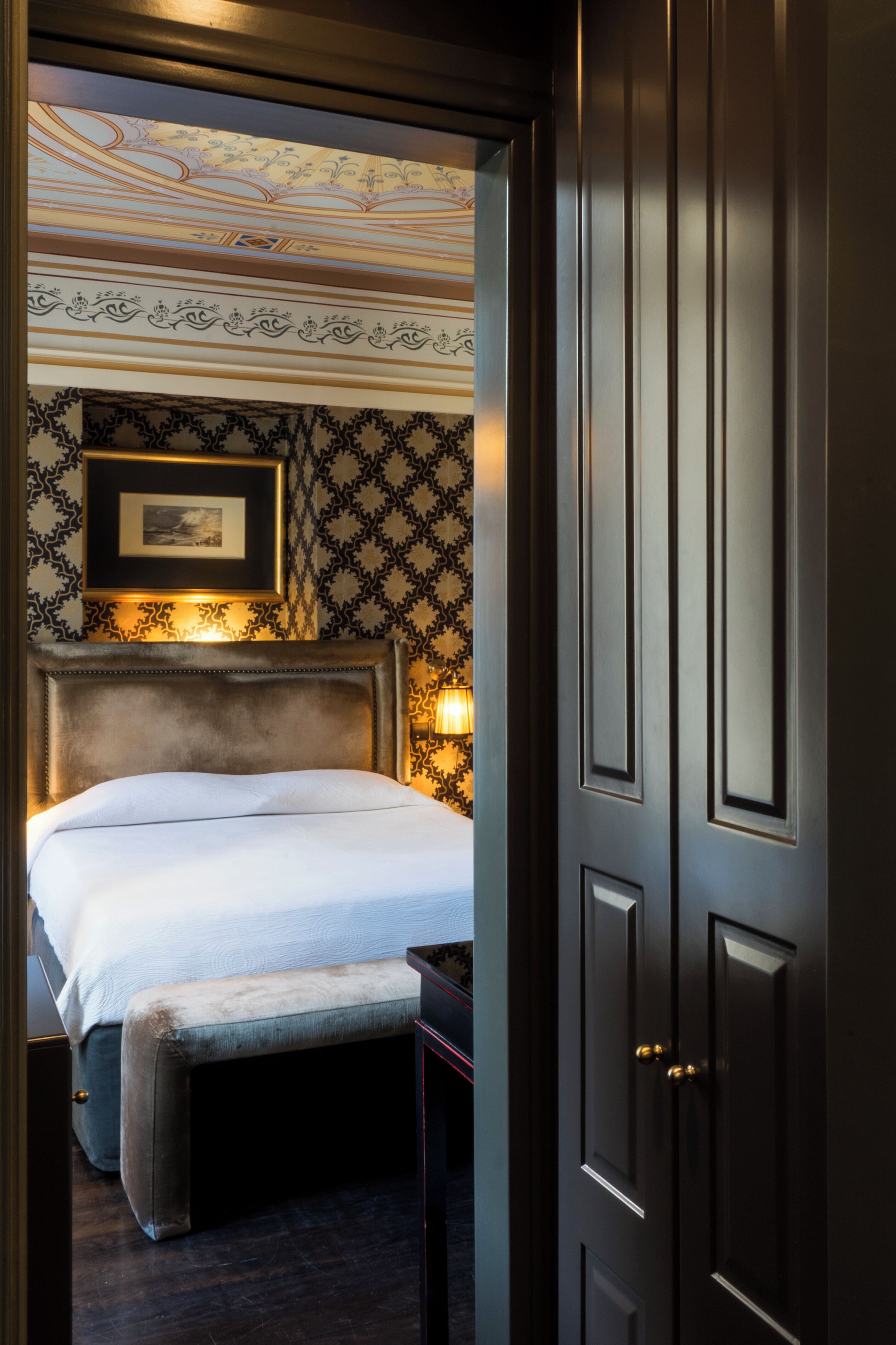 Habitación de hotel con una cama grande, un banco, un cuadro en la pared y un papel tapiz estampado.