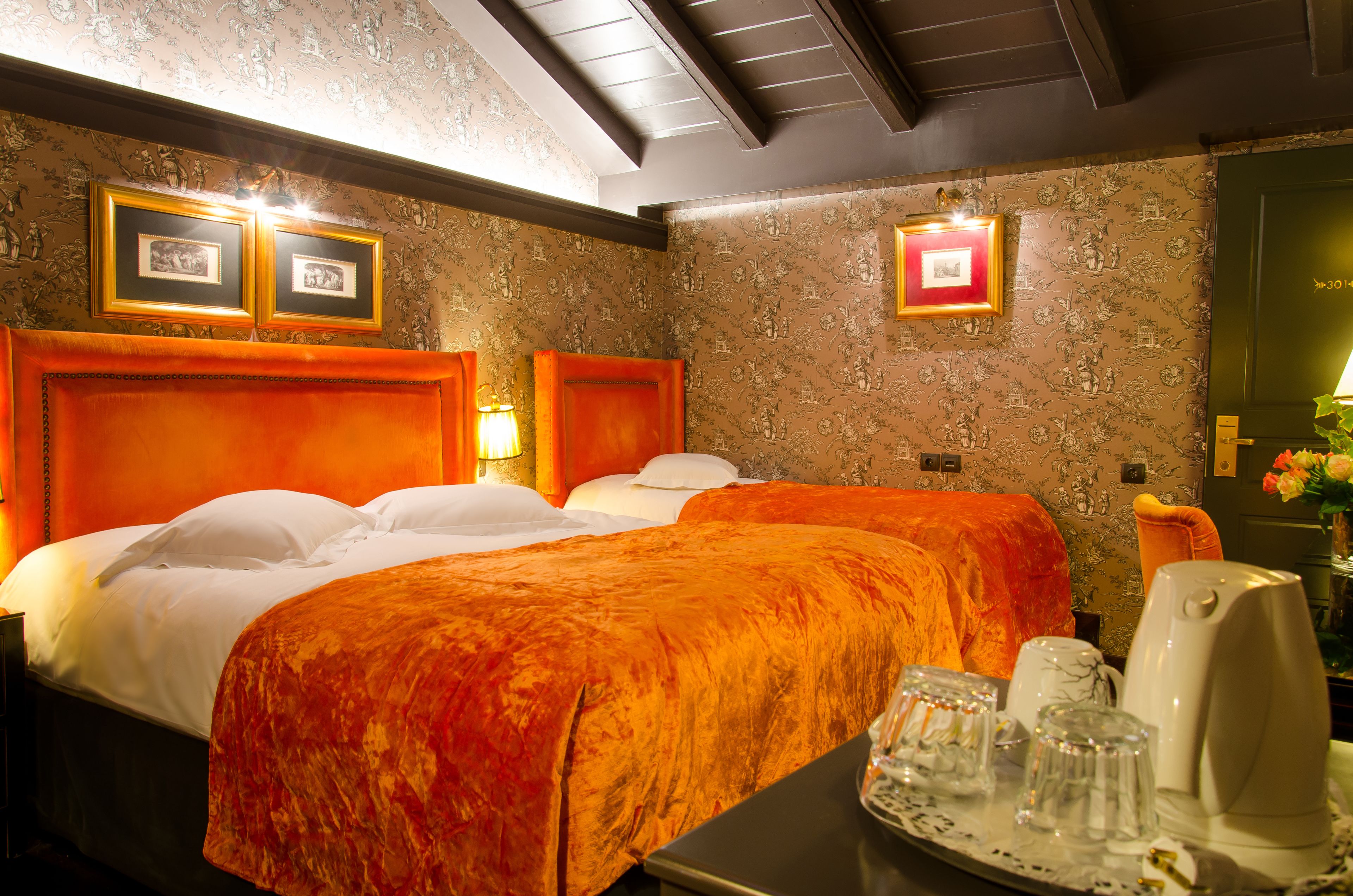 Une chambre d’hôtel avec deux lits, un plateau avec des verres et une bouilloire, et des tableaux encadrés accrochés au mur.