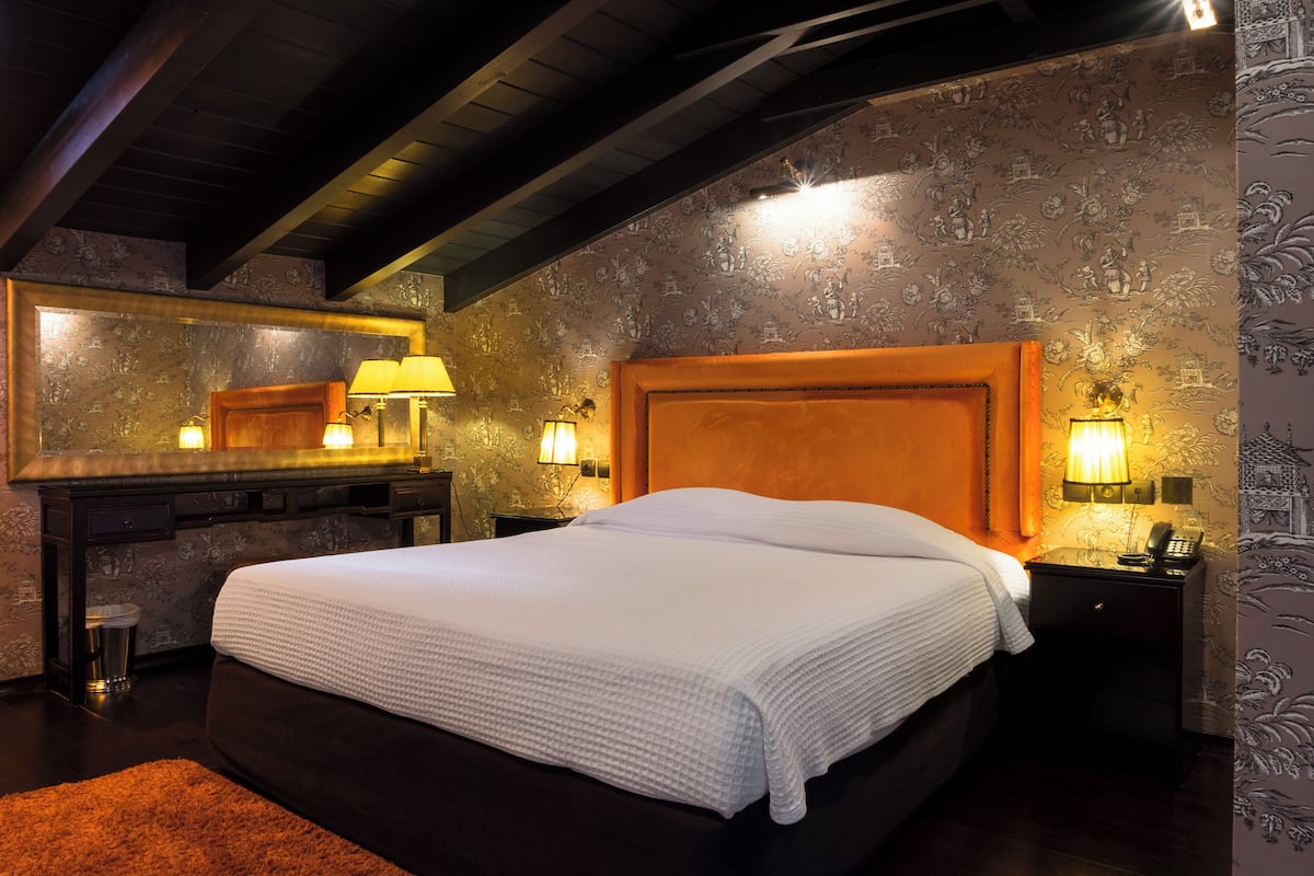 A hotel room with a large bed, a wooden headboard, a nightstand with a lamp, a mirror, and a desk with a lamp.