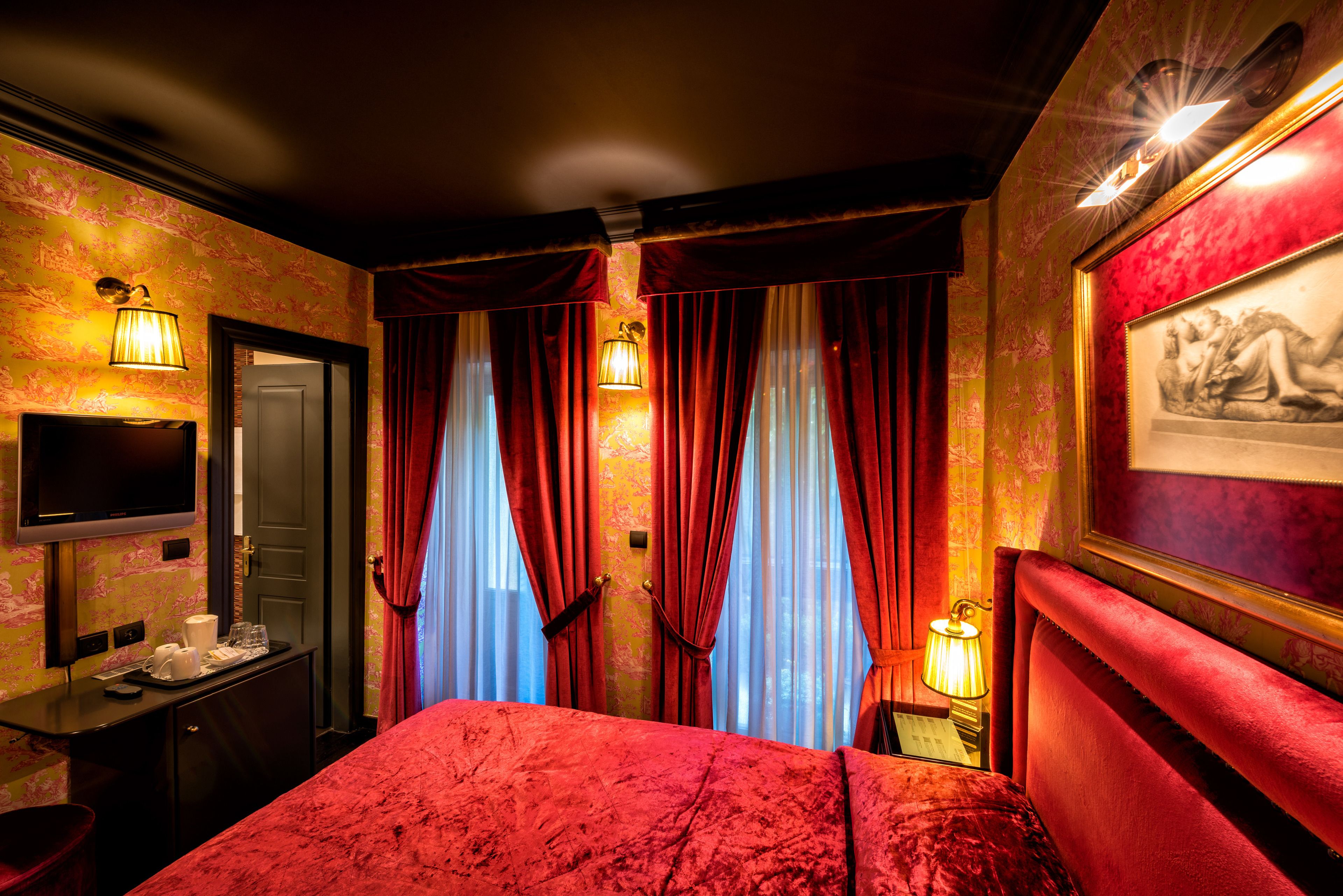 Een hotelkamer met een rode plaid, een bureau met een lamp, een televisie en een ingelijst schilderij aan de muur.