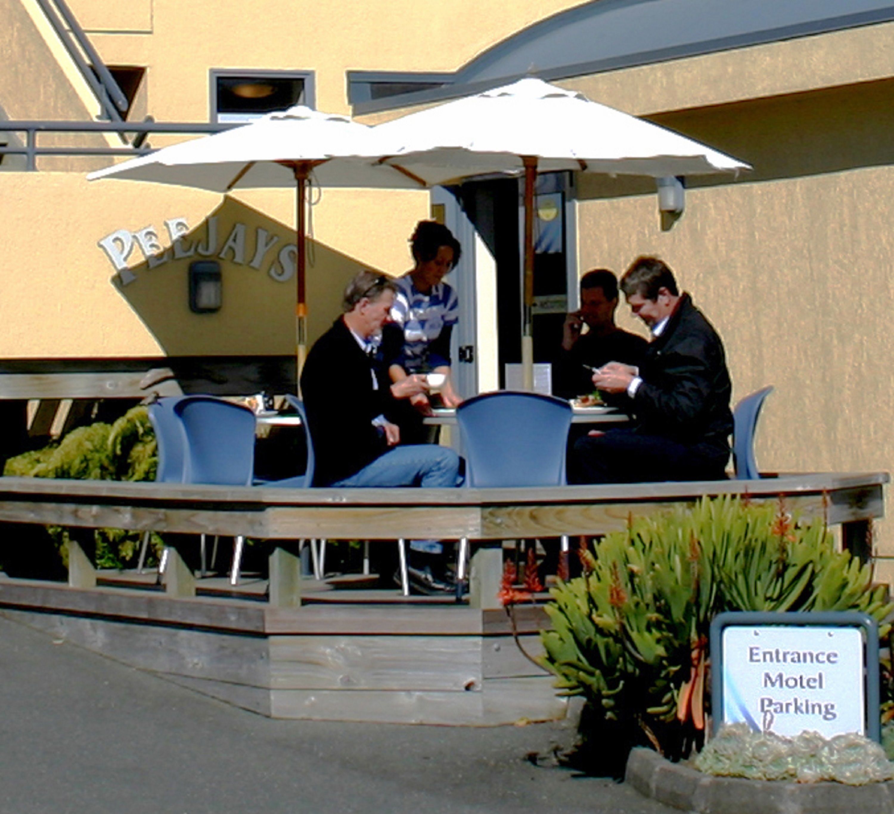 Un café al aire libre con mesas y sillas bajo sombrillas, un letrero que dice 'PEELAYS' y un cartel que indica 'Entrada, estacionamiento motel'.