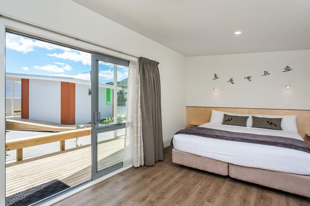 A modern hotel room with a large sliding glass door leading to an outdoor deck, a neatly made bed with a headboard, and wall-mounted light fixtures.