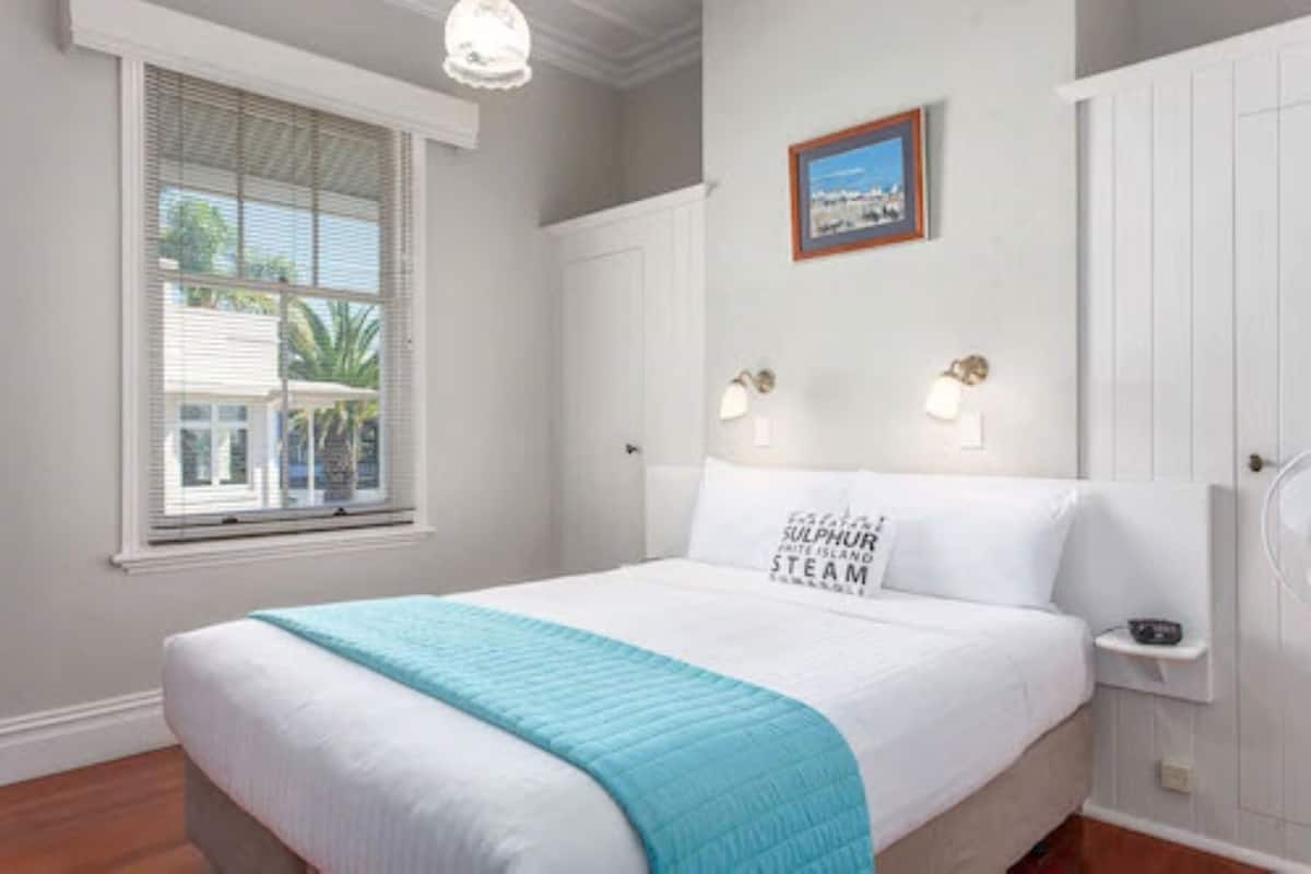 A neatly arranged bedroom with a bed, a window with blinds, a framed picture on the wall, and a nightstand with a lamp.