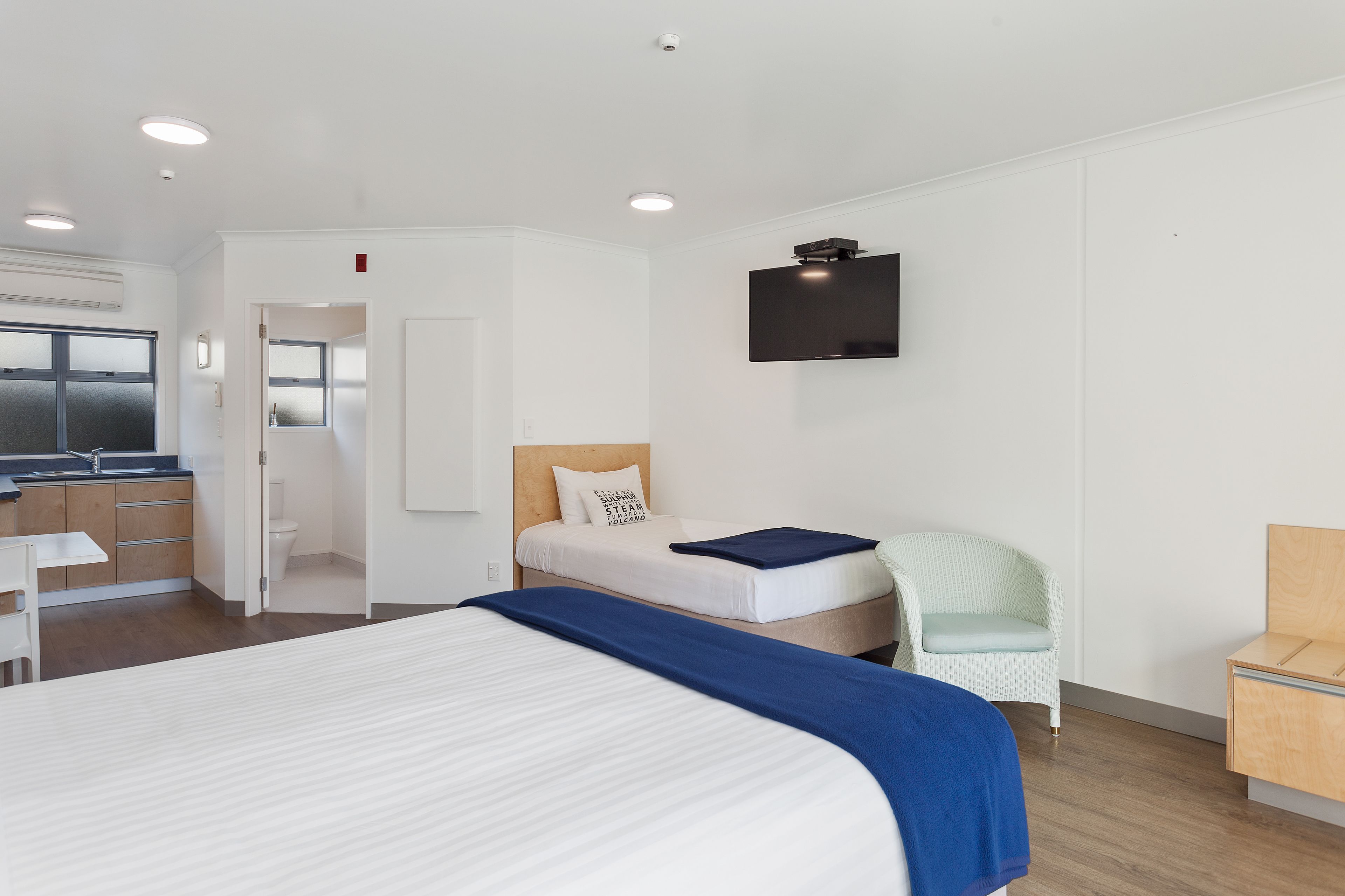 A modern hotel room with a large bed, a small desk, a chair, a TV, and a kitchenette.