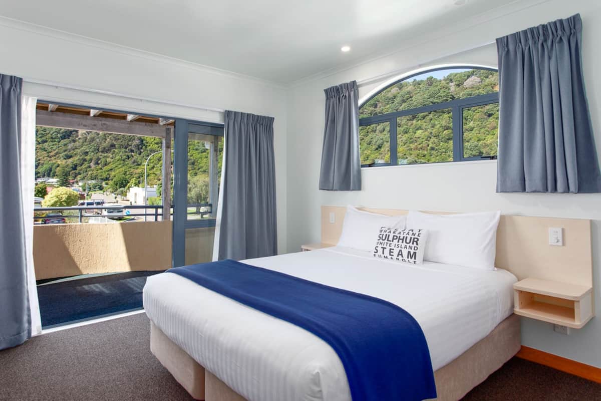 A hotel room with a large bed, a view of a green landscape, and a balcony with curtains.