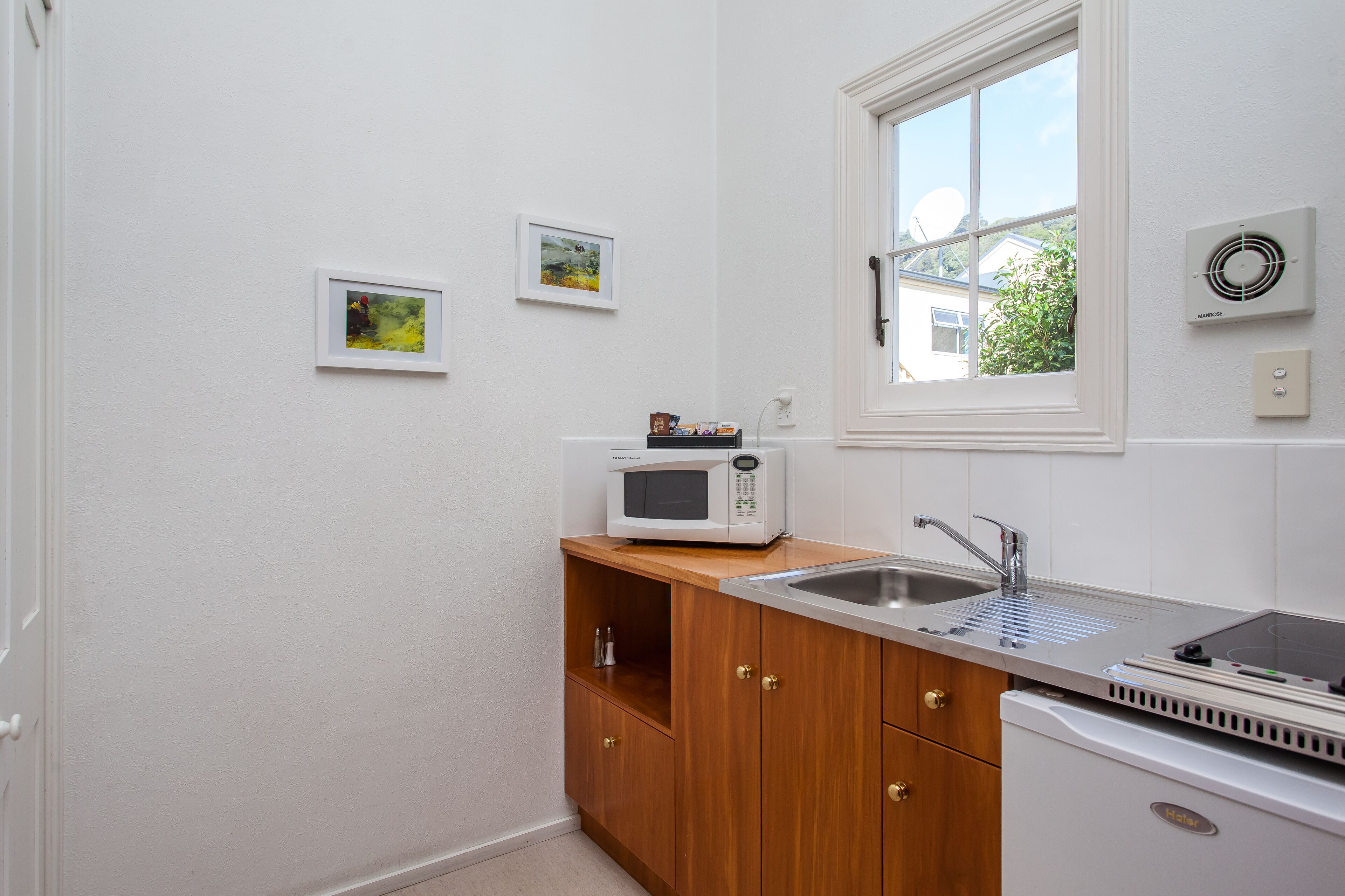Una cocina compacta con paredes blancas, un mueble de madera, un microondas, un lavamanos y una ventana con vistas a zonas verdes.