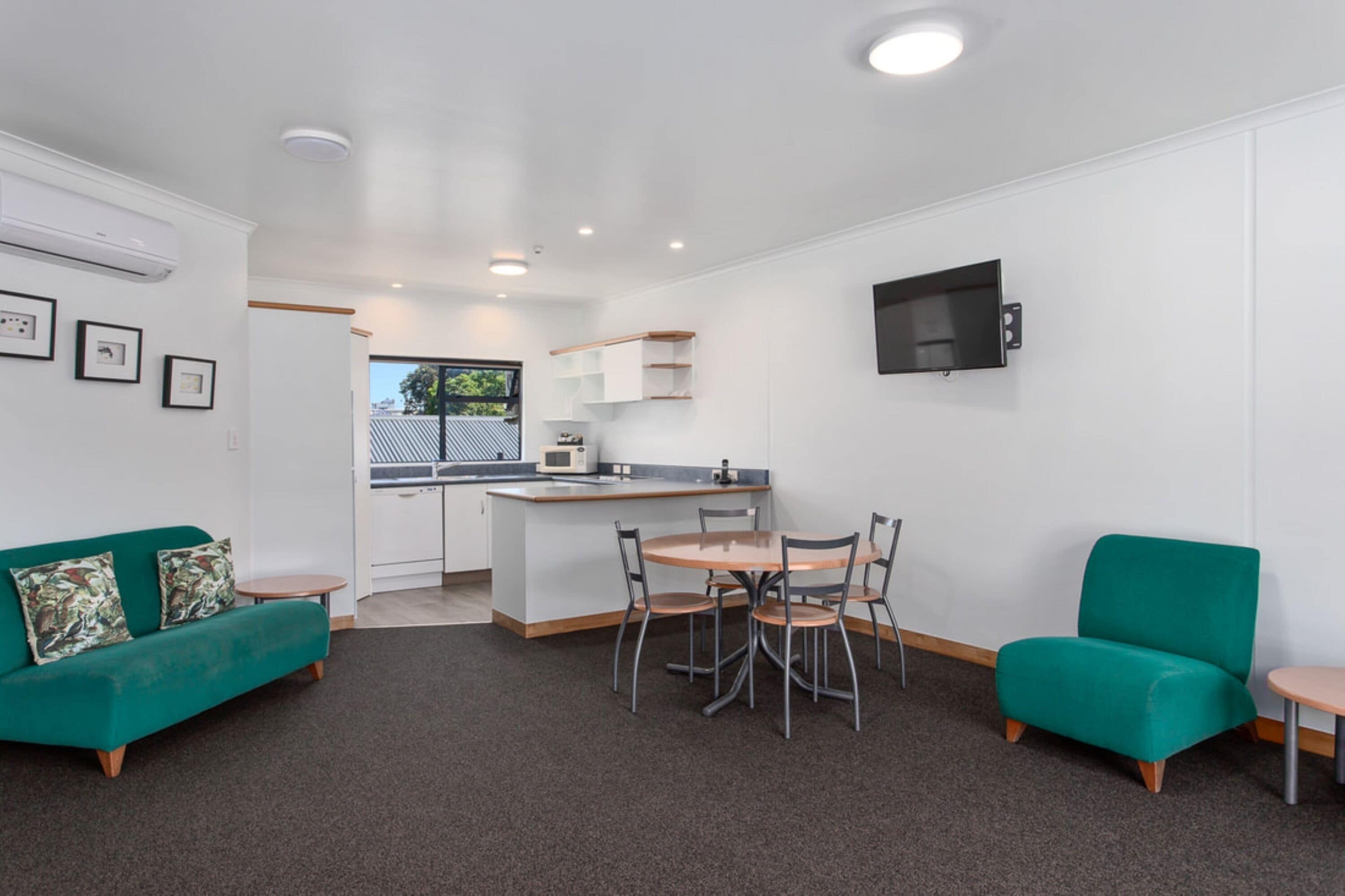 Una sala de estar moderna con un sofá verde, una mesa de comedor con sillas, una zona de cocina con microondas y un televisor montado en la pared.