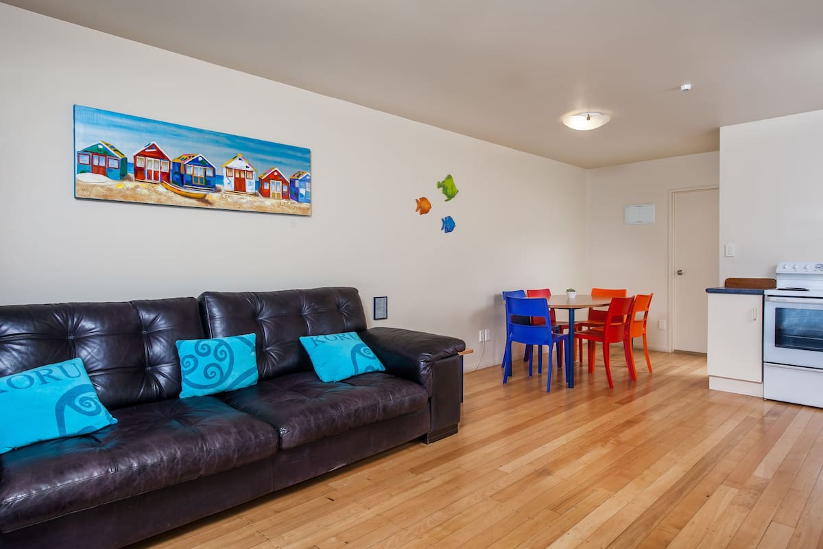 A living room with a brown leather sofa, a painting of beach houses, and a dining area with blue and red chairs.
