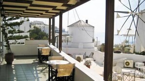 Una terraza en la azotea con mobiliario exterior, un edificio blanco al fondo y vistas al mar.