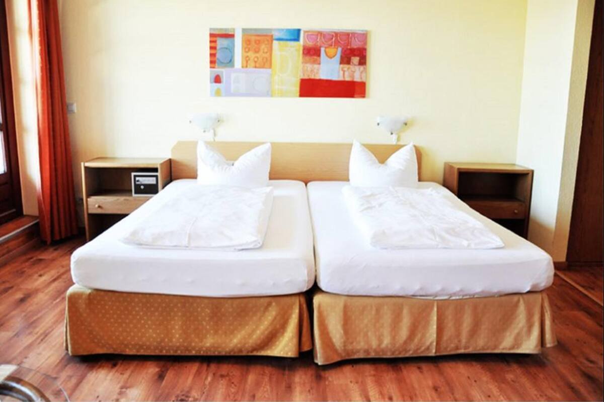 A hotel room with two single beds, wooden flooring, a nightstand with a lamp, and a wall with colorful artwork.