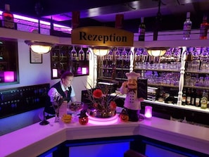 A bar area with a 'Rezeption' sign, shelves with bottles, and a decorative centerpiece with pumpkins and a chef figure.