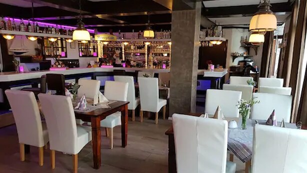 A restaurant with white chairs and tables, a bar area with shelves of bottles, and pendant lighting. - Hotel an der Hörn (Kiel)