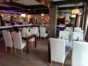 A restaurant with white chairs and tables, a bar area with shelves of bottles, and pendant lighting. - Hotel an der Hörn (Kiel)