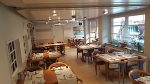 A dining area with multiple tables set for service, wooden chairs, and a large window with a view. - Hotel an der Hörn (Kiel)